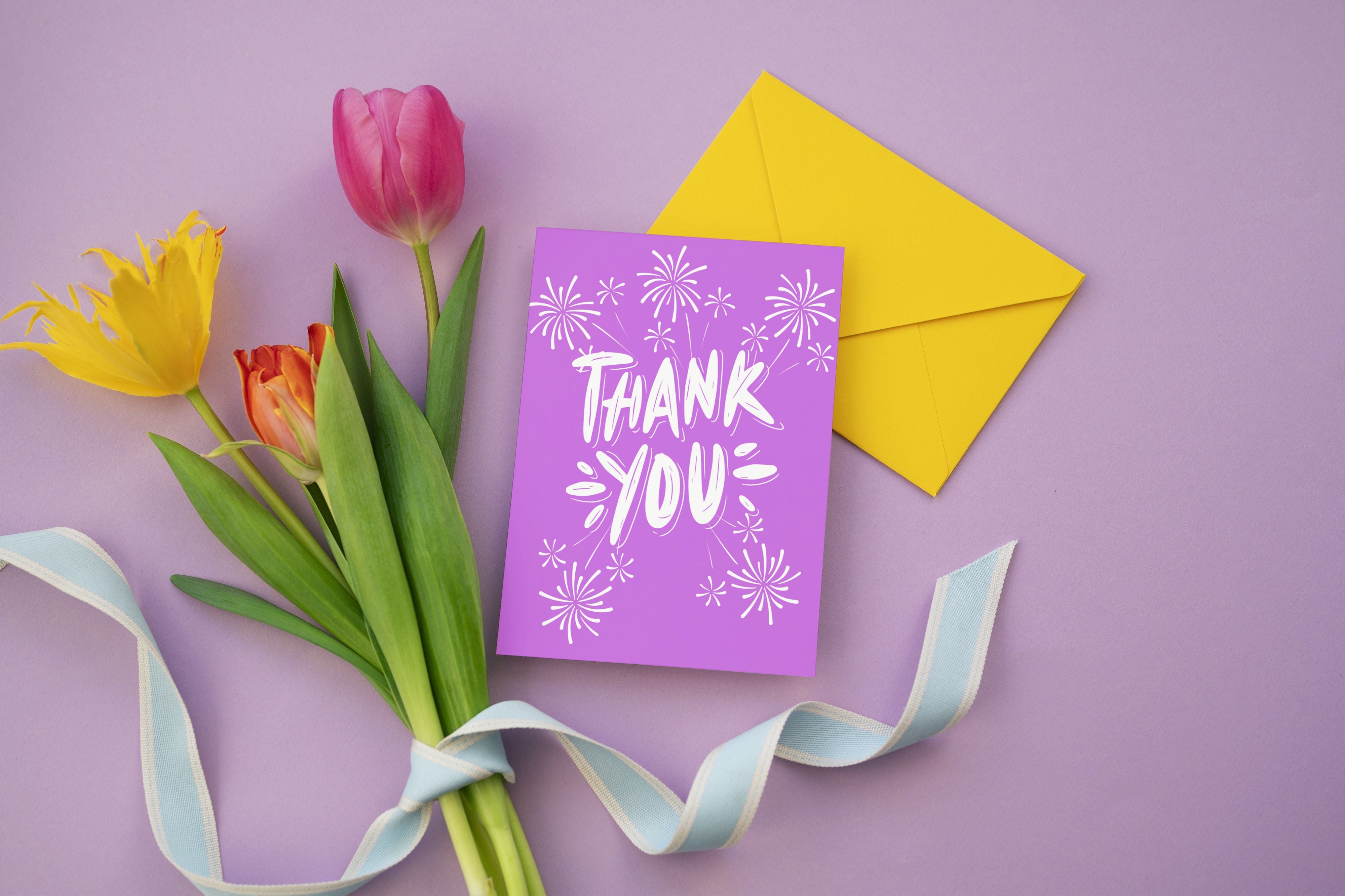 Purple card saying thank you with fireworks sitting on purple table with flowers next to it.