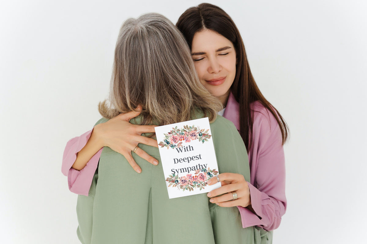 Young woman hugging older woman, holding floral card saying With deepest sympathy.